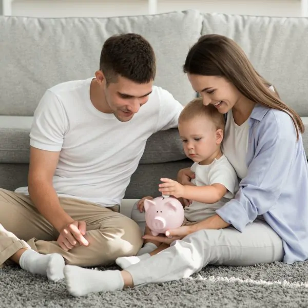 Family Budget. Happy Parents Looking At Infant Son Putting Coins To Piggybank